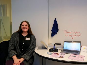 Chloe Castille sitting by her laptop with her project, "Michigan Voices Podcast, Season 7 Episode 9 The Case for Community: Why Community College is Vital to Everyone, even U-M Students."